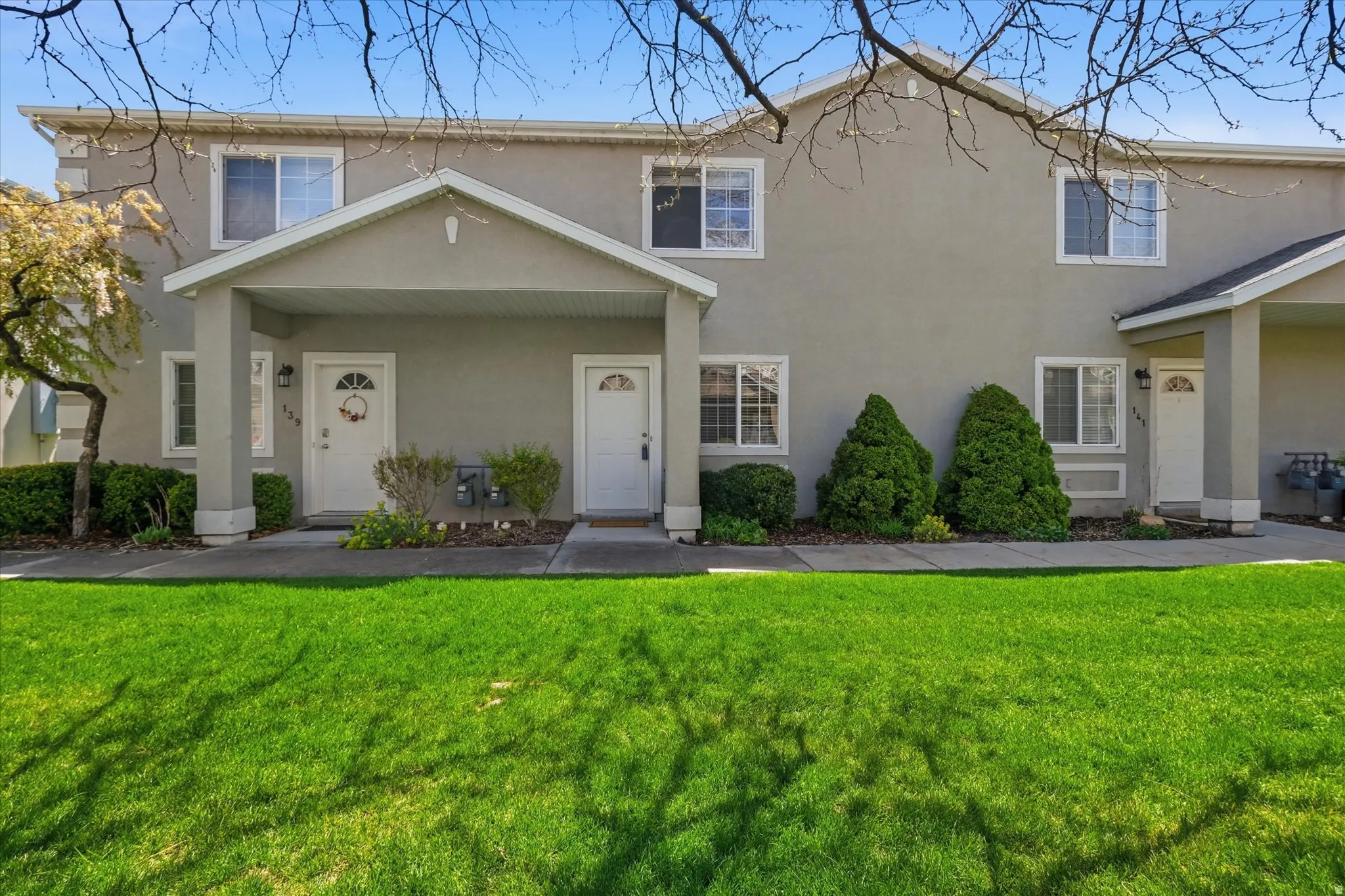 Stucco exterior townhome featuring a covered entry, white-framed windows, and manicured front landscaping