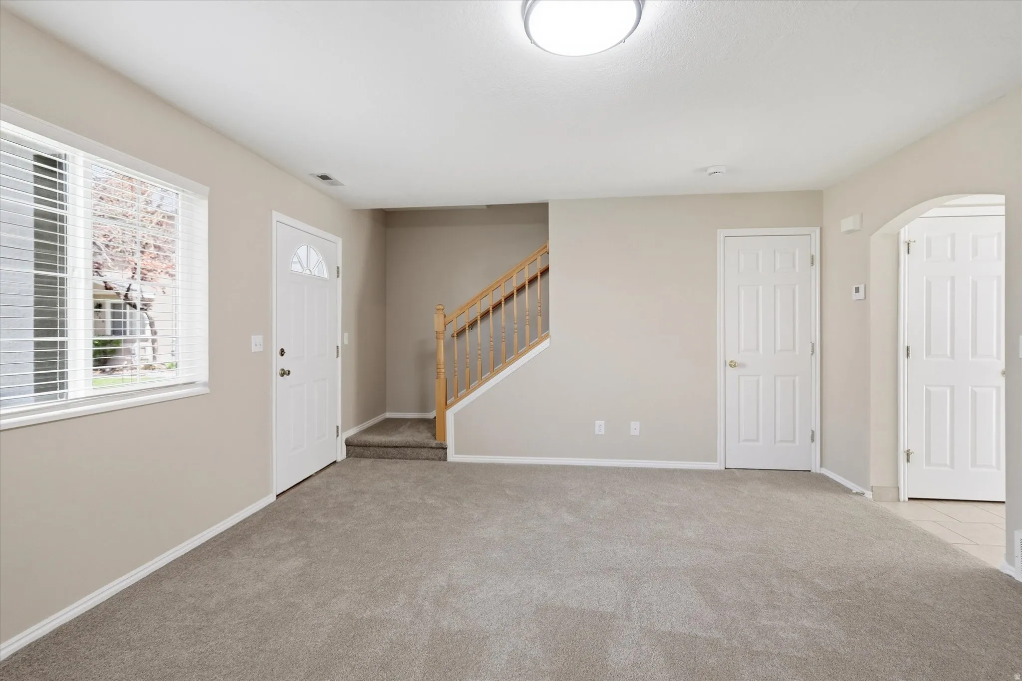 Spacious entry area featuring light gray carpeting, neutral wall tones, a white paneled entry door with an arched window, and a wood-railed staircase