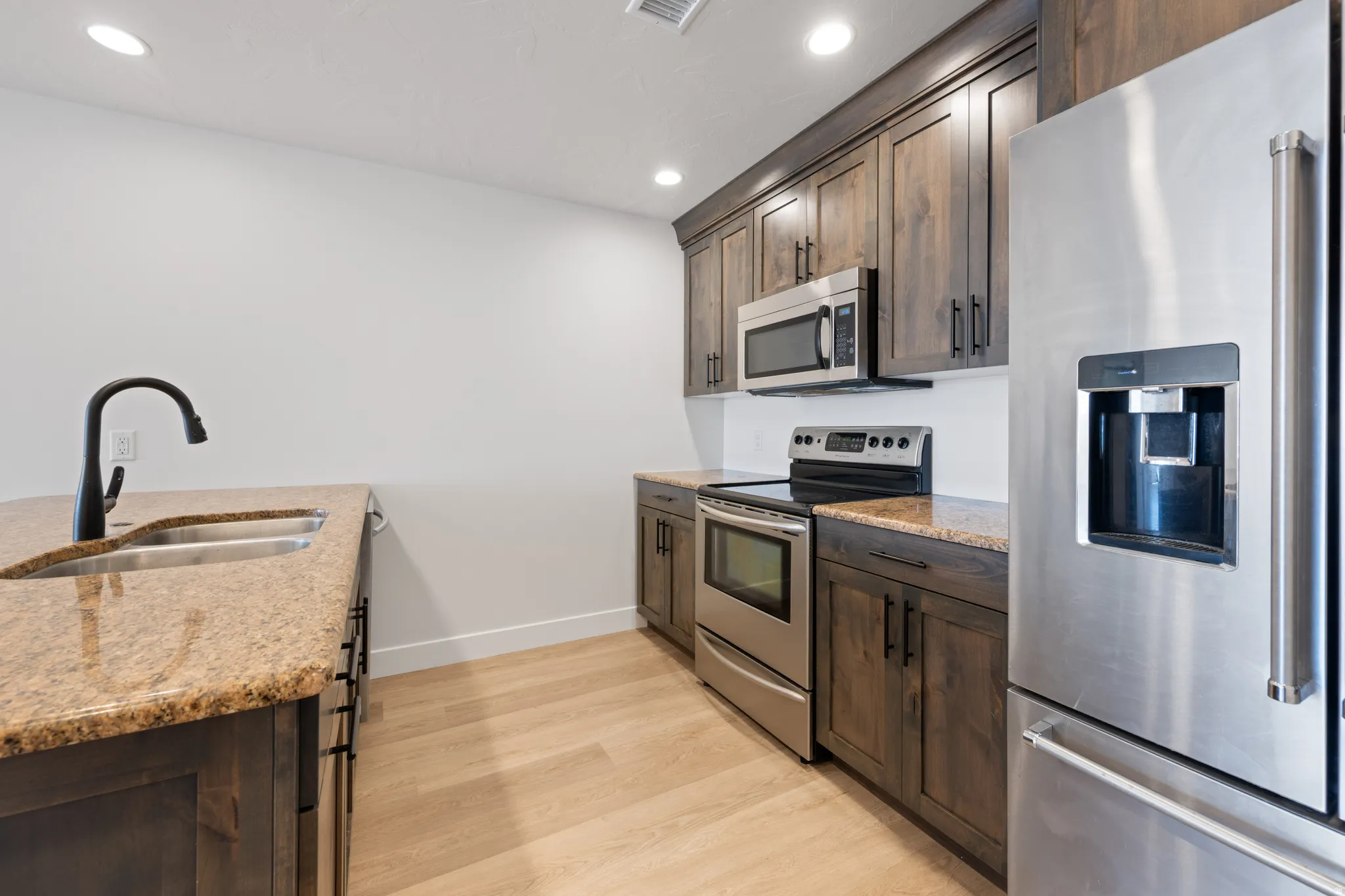 Kitchen with stainless steel appliances, dark wood finish cabinetry, light wood-type flooring, light stone countertops, and recessed lighting