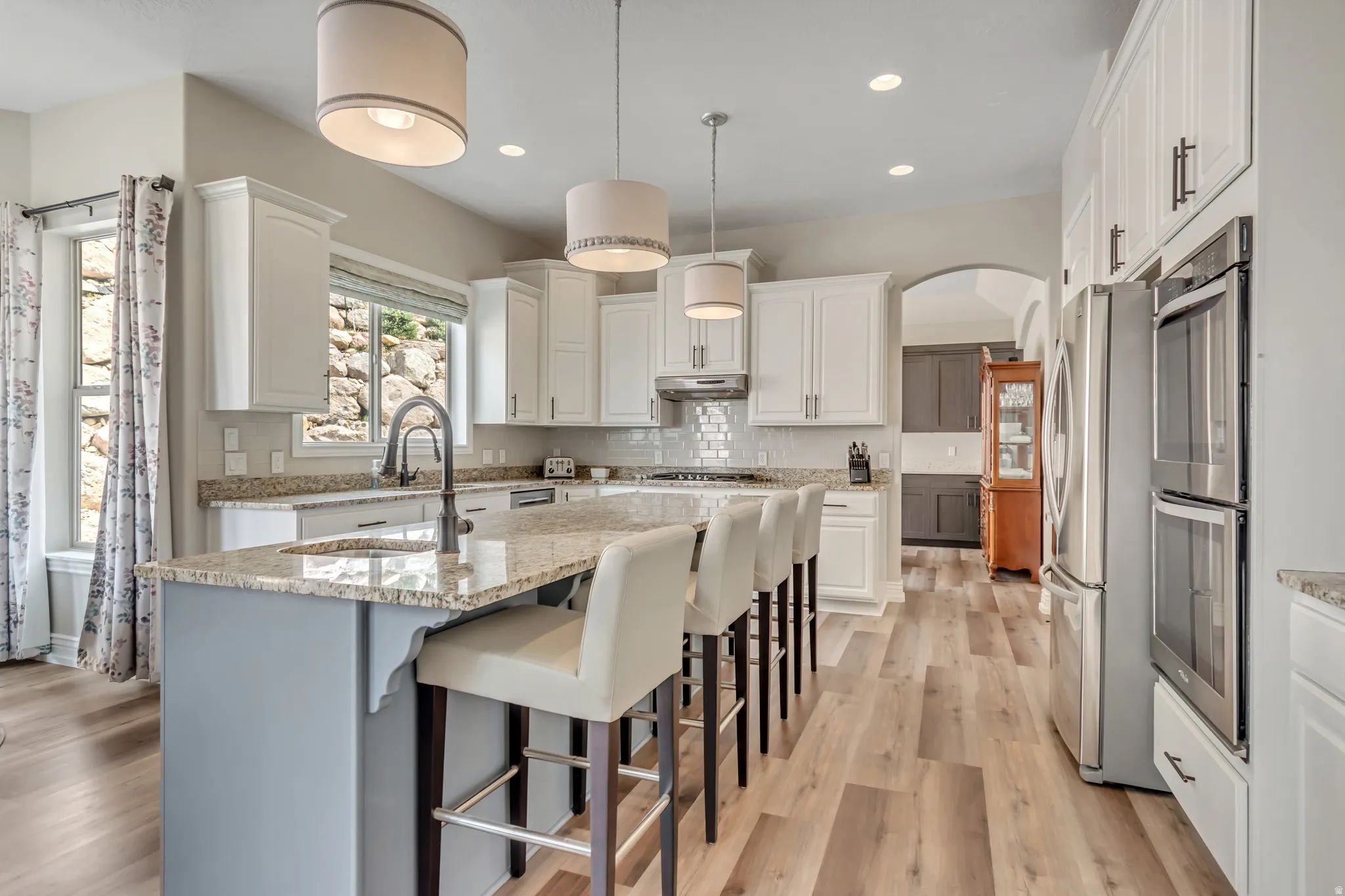 Two tone kitchen with light stone countertops, a center island with sink, a kitchen bar, stainless steel appliances, and pendant lighting