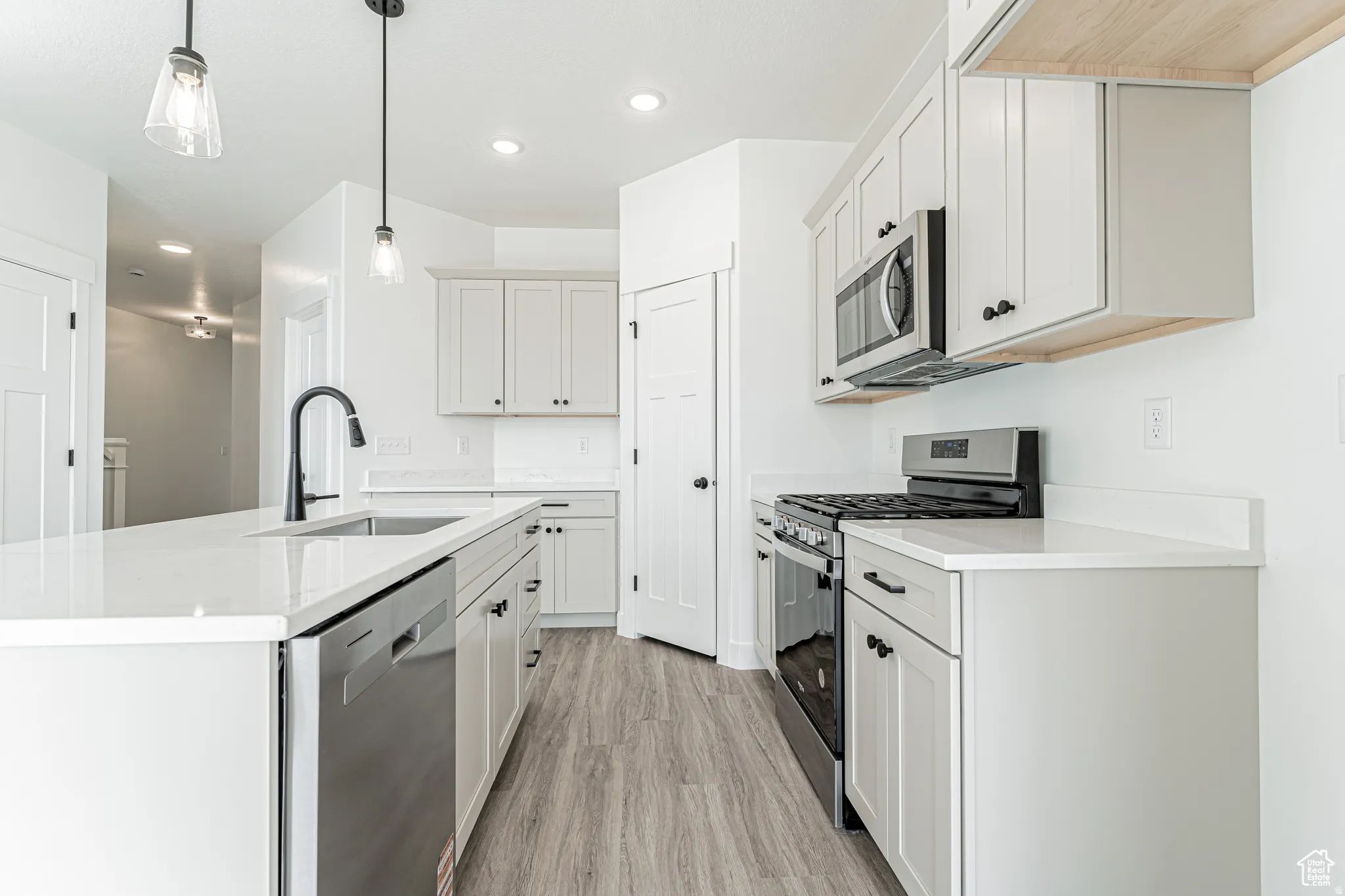 Kitchen featuring stainless steel appliances, light wood-type flooring, an island with sink, pendant lighting, and light stone countertops