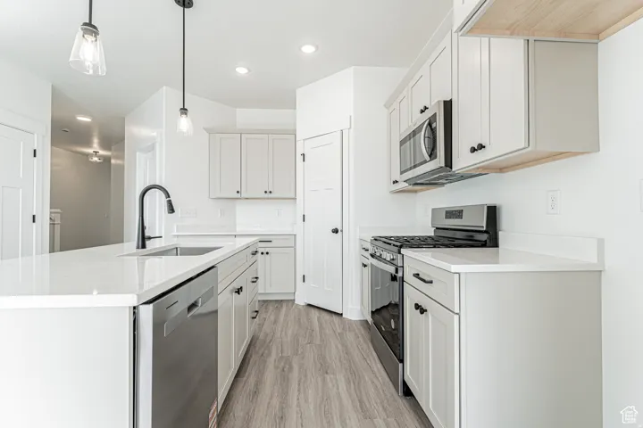 Kitchen featuring stainless steel appliances, light wood-type flooring, an island with sink, pendant lighting, and light stone countertops