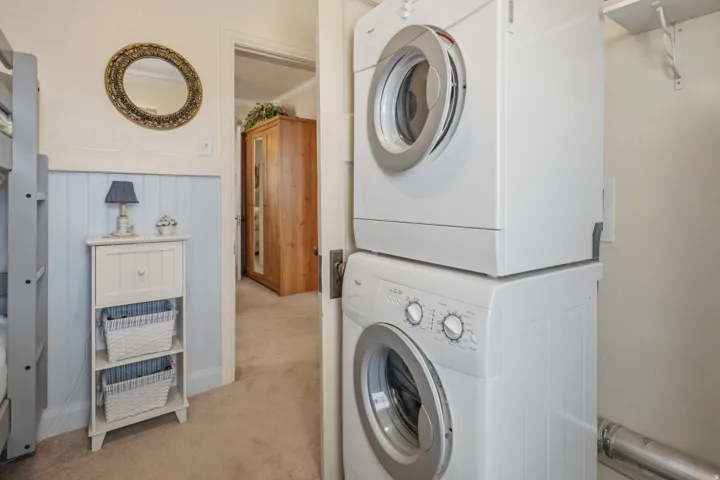Laundry area featuring light colored carpet, ornamental molding, stacked washing machine and dryer, and wainscoting