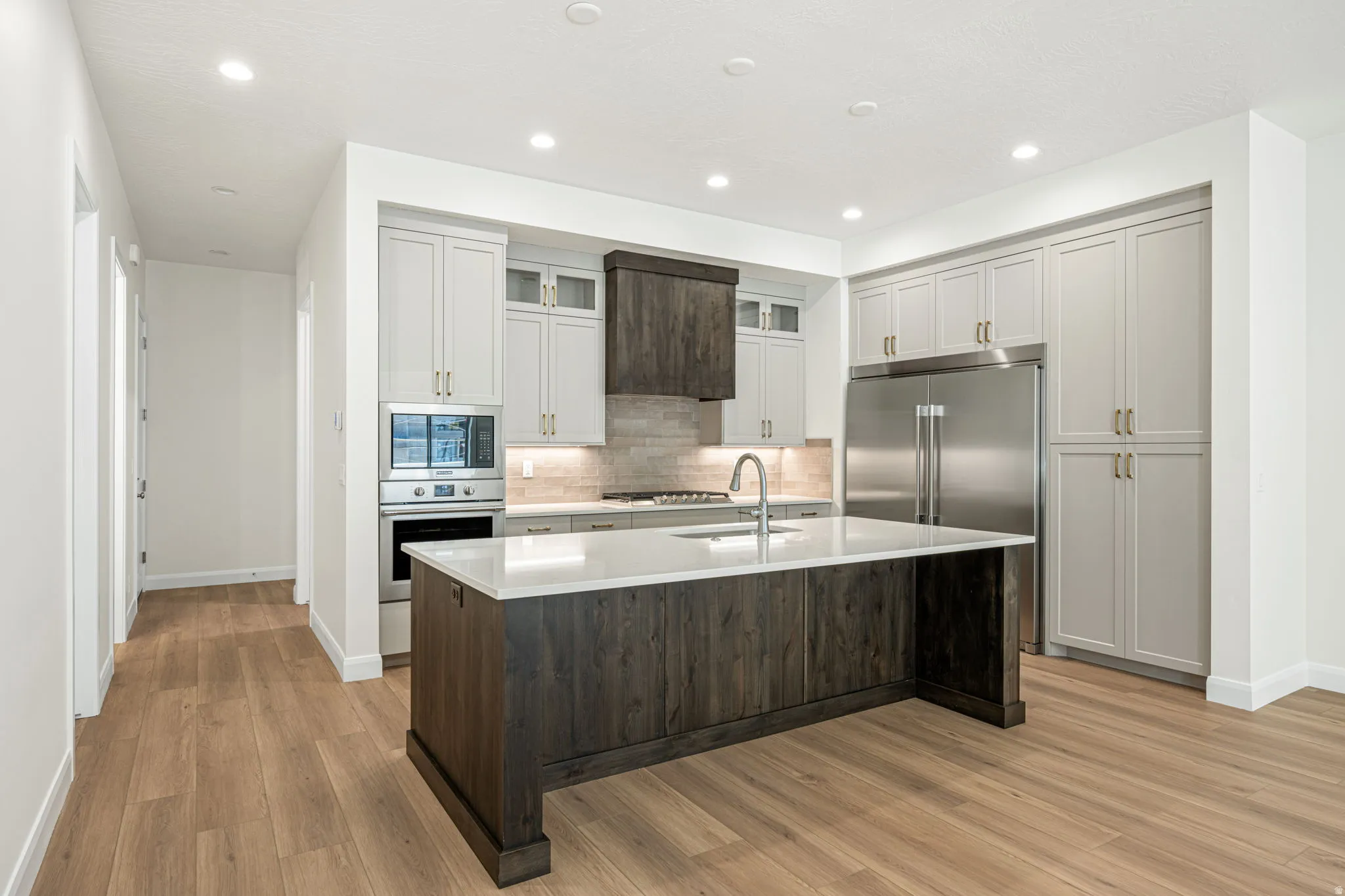 Modern kitchen featuring a large central island with a light-toned countertop, wood-finish flooring, recessed lighting, stainless steel appliances, and two-tone cabinetry with gold-finish hardware