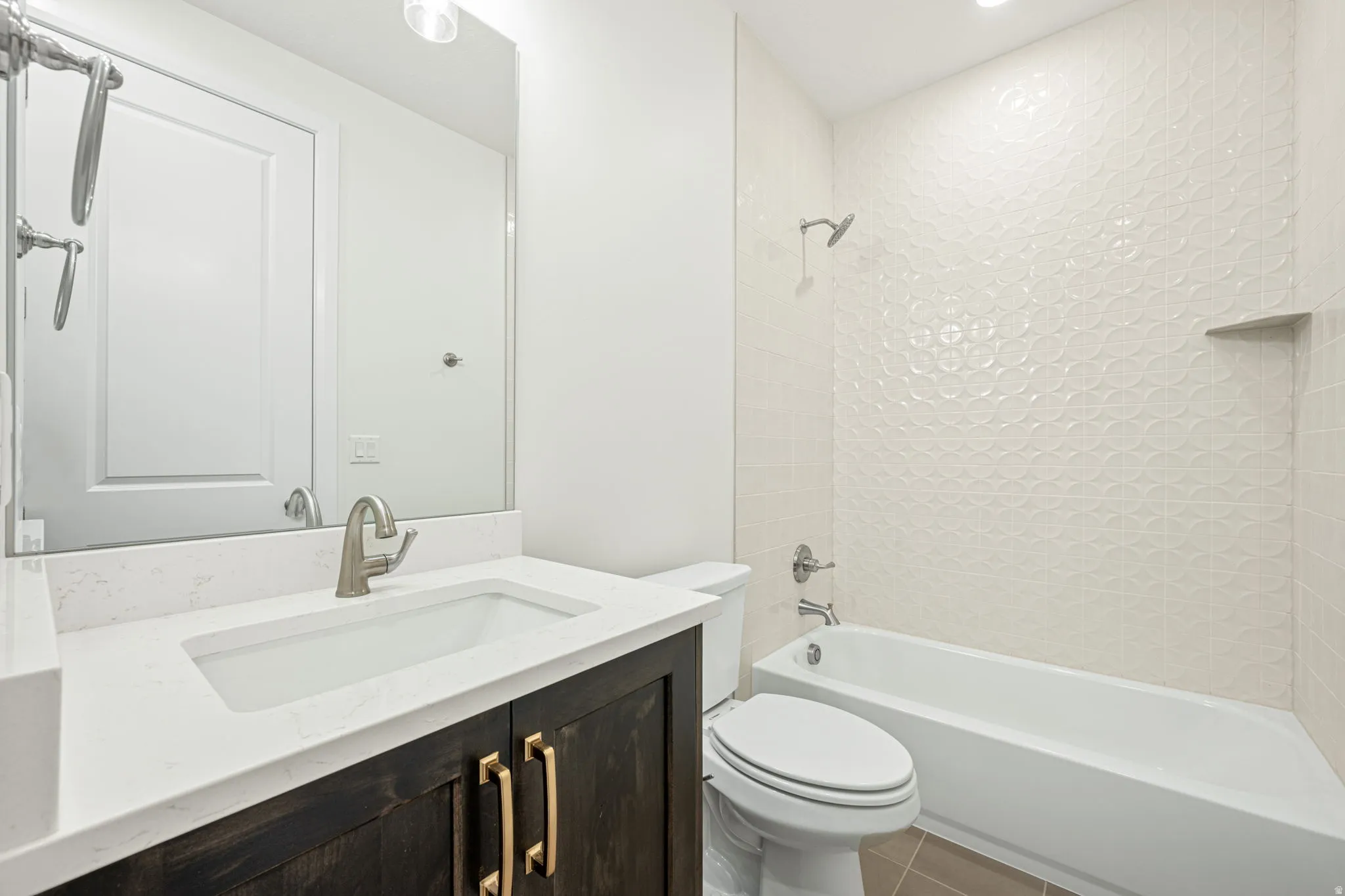 Bathroom featuring a vanity with a white countertop and rectangular undermount sink, dark wood-finish cabinetry with brass-tone hardware, a full-width mirror, and a tub-shower combination with textured white wall tile and an integrated niche