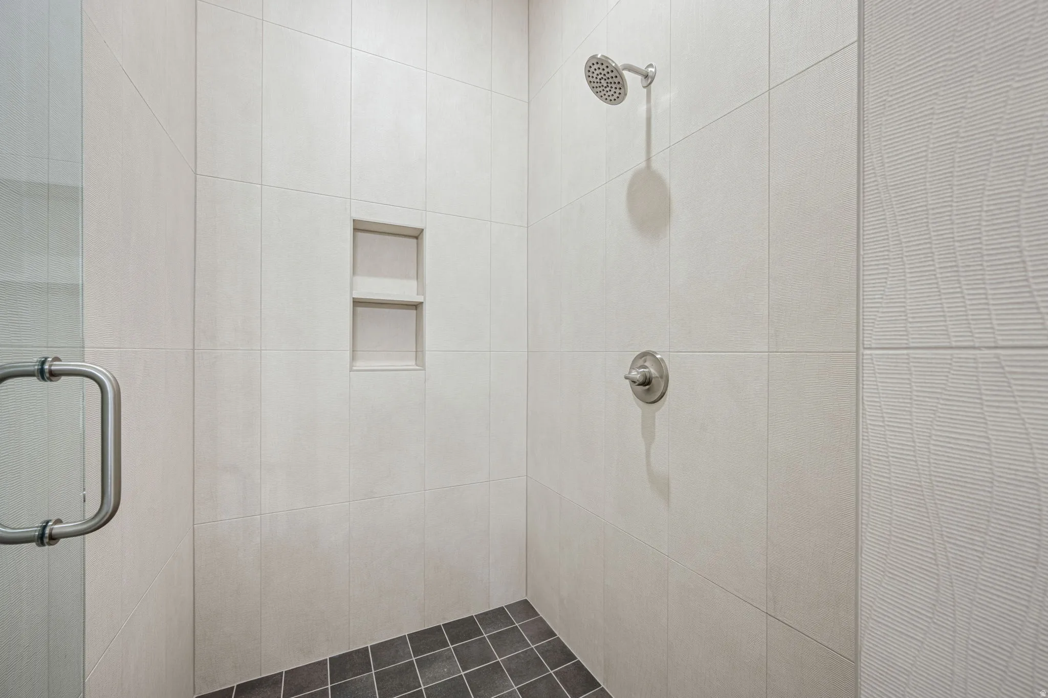 Walk-in shower with light-toned rectangular wall tiles, a built-in shower niche, a brushed nickel shower head and control, and dark square floor tiles