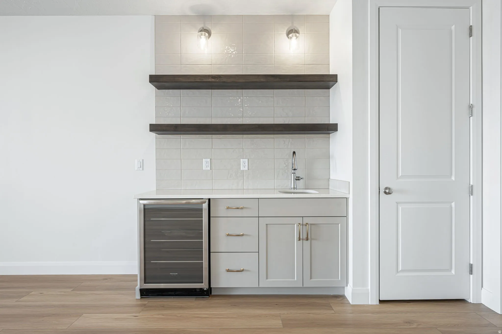 Built-in wet bar featuring light grey cabinetry, a stainless steel sink with a chrome faucet, a beverage refrigerator, and open wood shelving