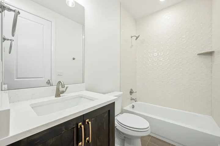 Bathroom featuring a vanity with a white countertop and rectangular undermount sink, dark wood-finish cabinetry with brass-tone hardware, a full-width mirror, and a tub-shower combination with textured white wall tile and an integrated niche