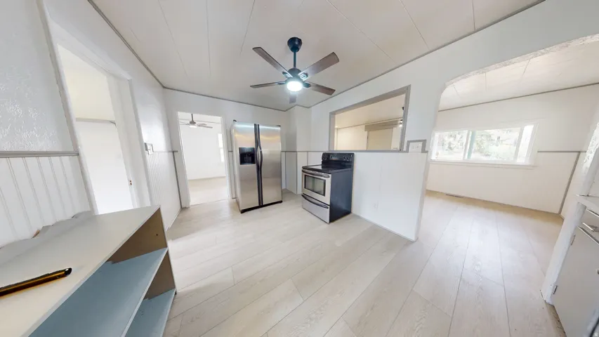 Bright kitchen space featuring light wood-finish flooring, stainless steel appliances, a ceiling fan with integrated lighting, white beadboard wainscoting, and an archway opening to an additional room