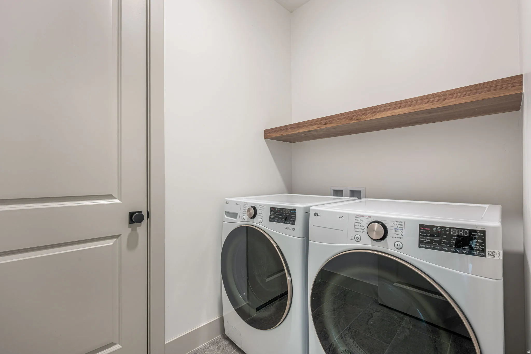 Dedicated laundry space featuring a floating wood-finish shelf, light gray wall paneling, and dark gray flooring