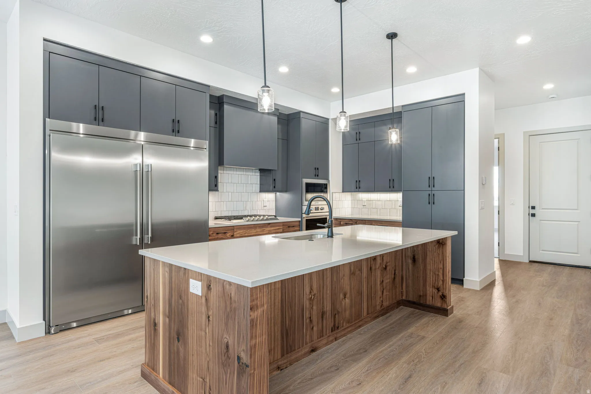 Modern kitchen featuring a large wood-finish island with a light-toned countertop, sleek dark gray cabinetry, stainless steel appliances, and a tiled backsplash