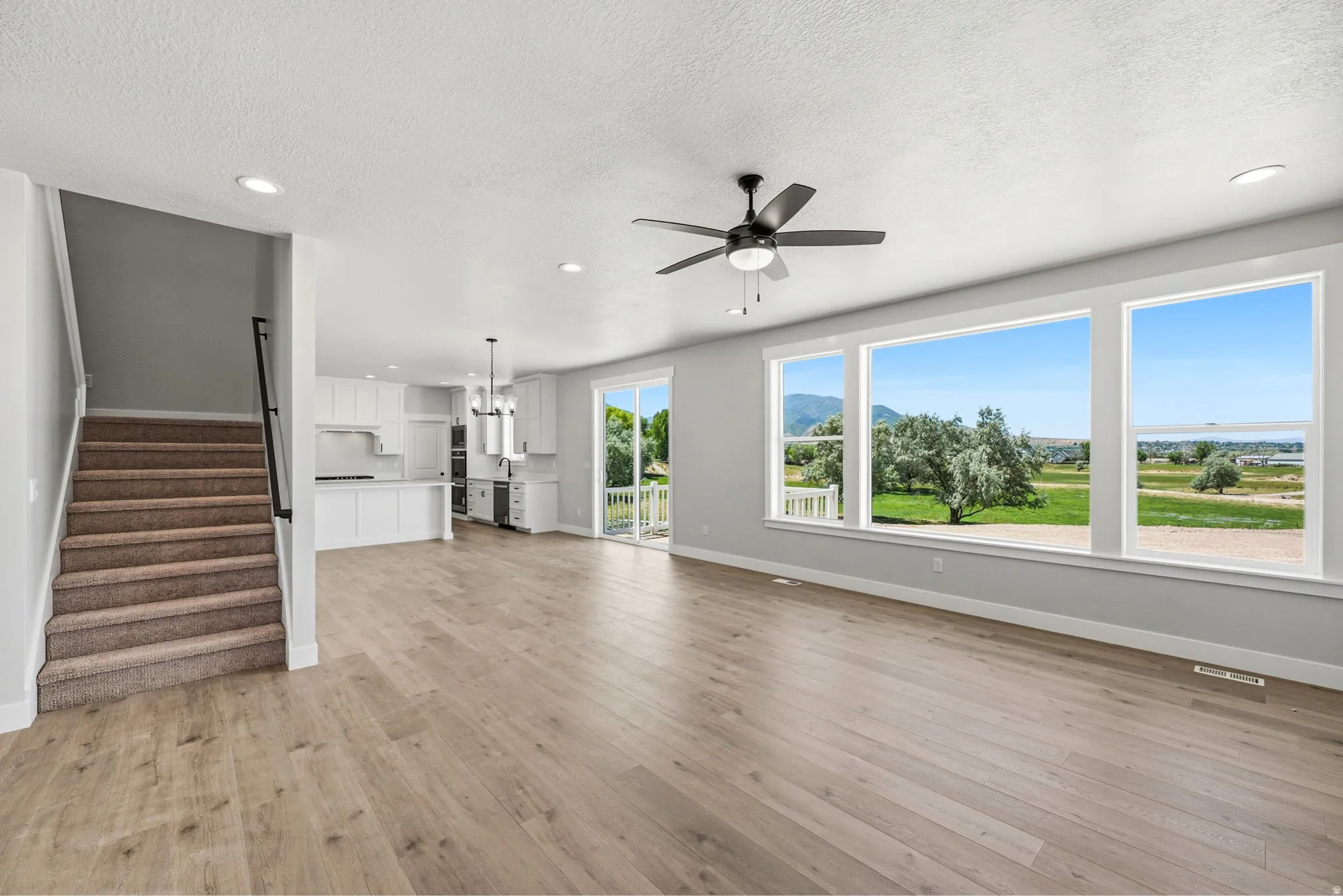 Spacious open-concept living area featuring wood-finish flooring, a contemporary ceiling fan, and large windows with surrounding scenery