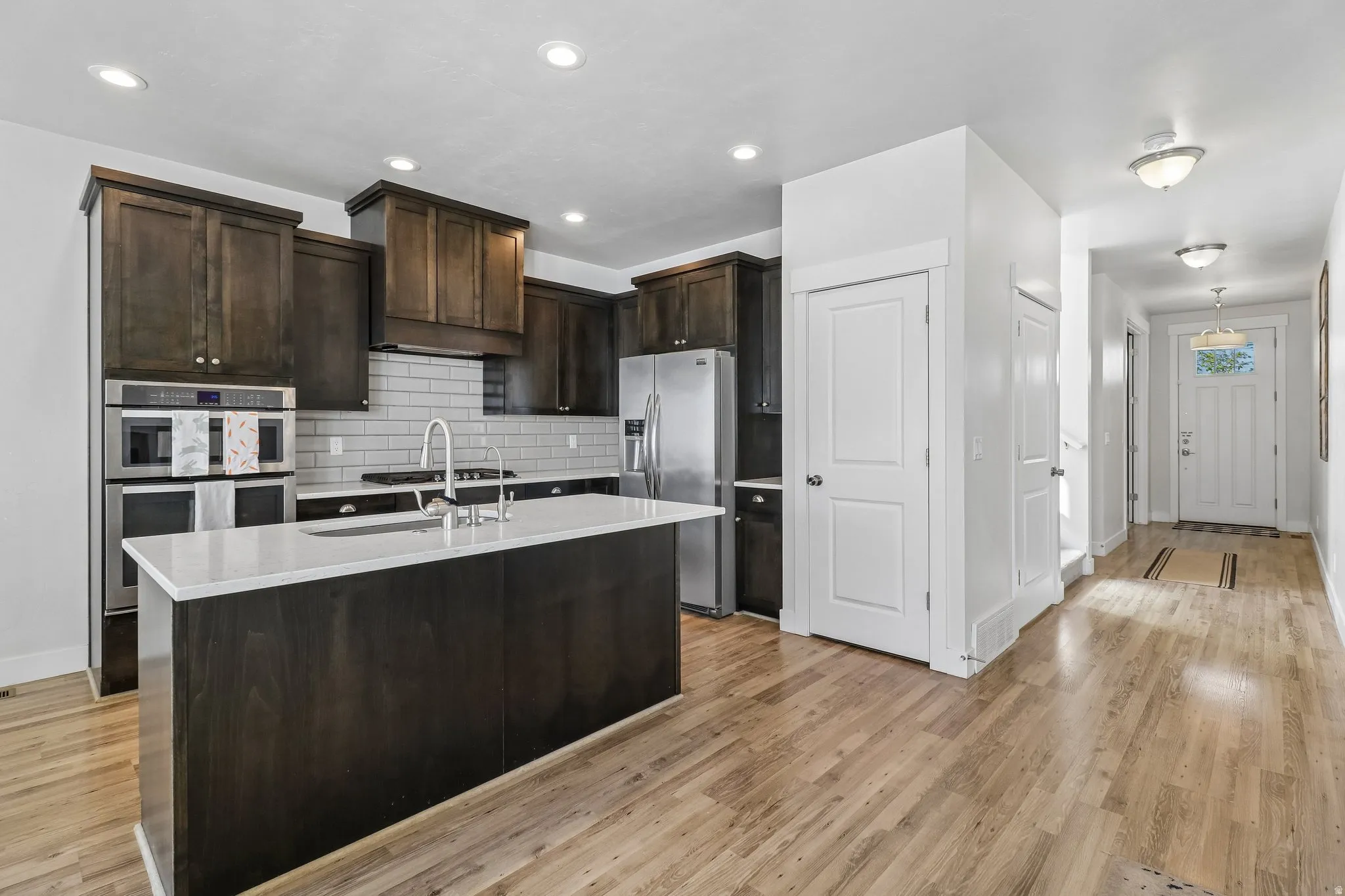 Kitchen featuring dark wood finish cabinets, stainless steel appliances, light stone counters, a center island with sink, and light wood finished floors