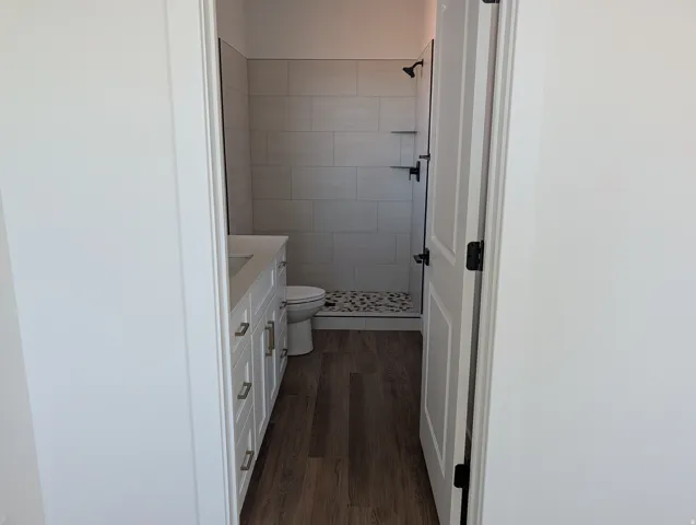 Bathroom featuring wood-finish flooring, a double vanity with white cabinetry and integrated sinks, and a walk-in shower with large format tile walls and a pebble-finish floor