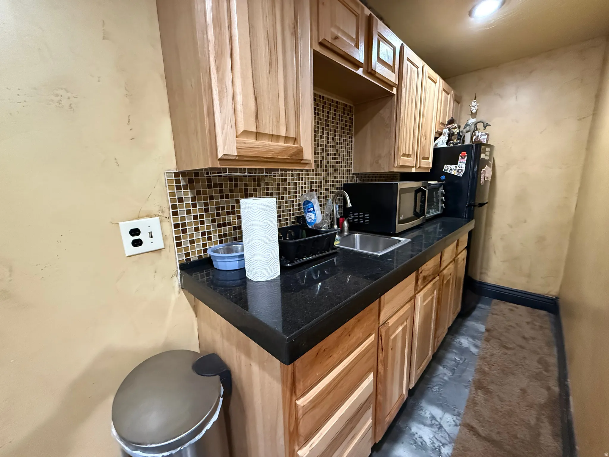Kitchen featuring dark countertops, light wood finish cabinetry, backsplash, freestanding refrigerator, and stainless steel microwave