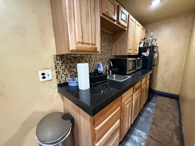 Kitchen featuring dark countertops, light wood finish cabinetry, backsplash, freestanding refrigerator, and stainless steel microwave