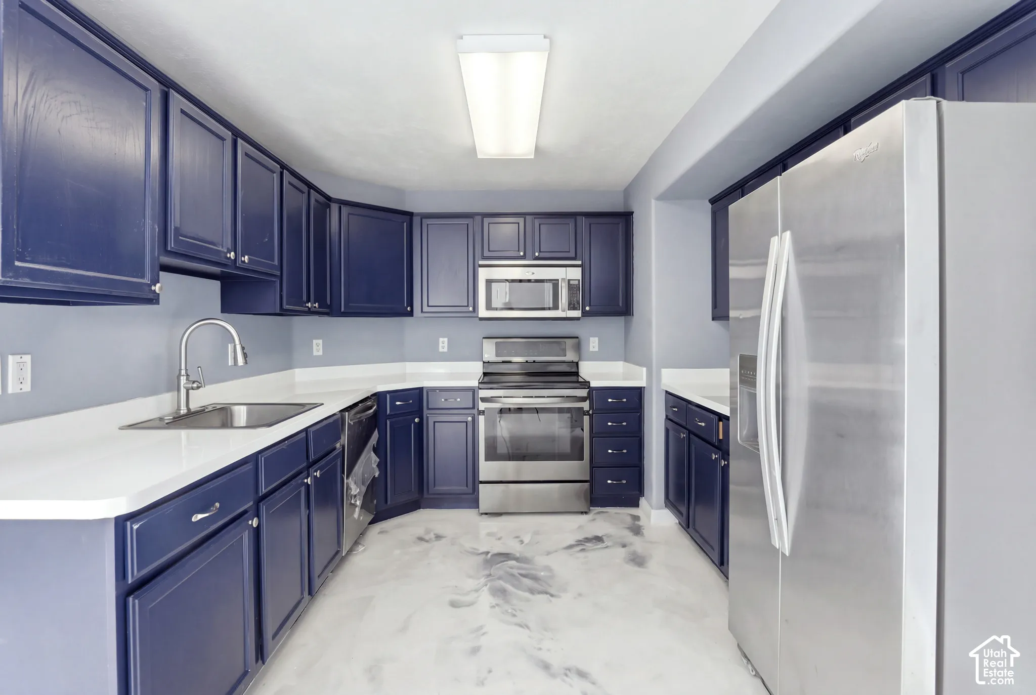 Kitchen featuring deep blue shaker-style cabinetry, white solid-surface countertops, stainless steel appliances, and polished flooring with a decorative marbling finish