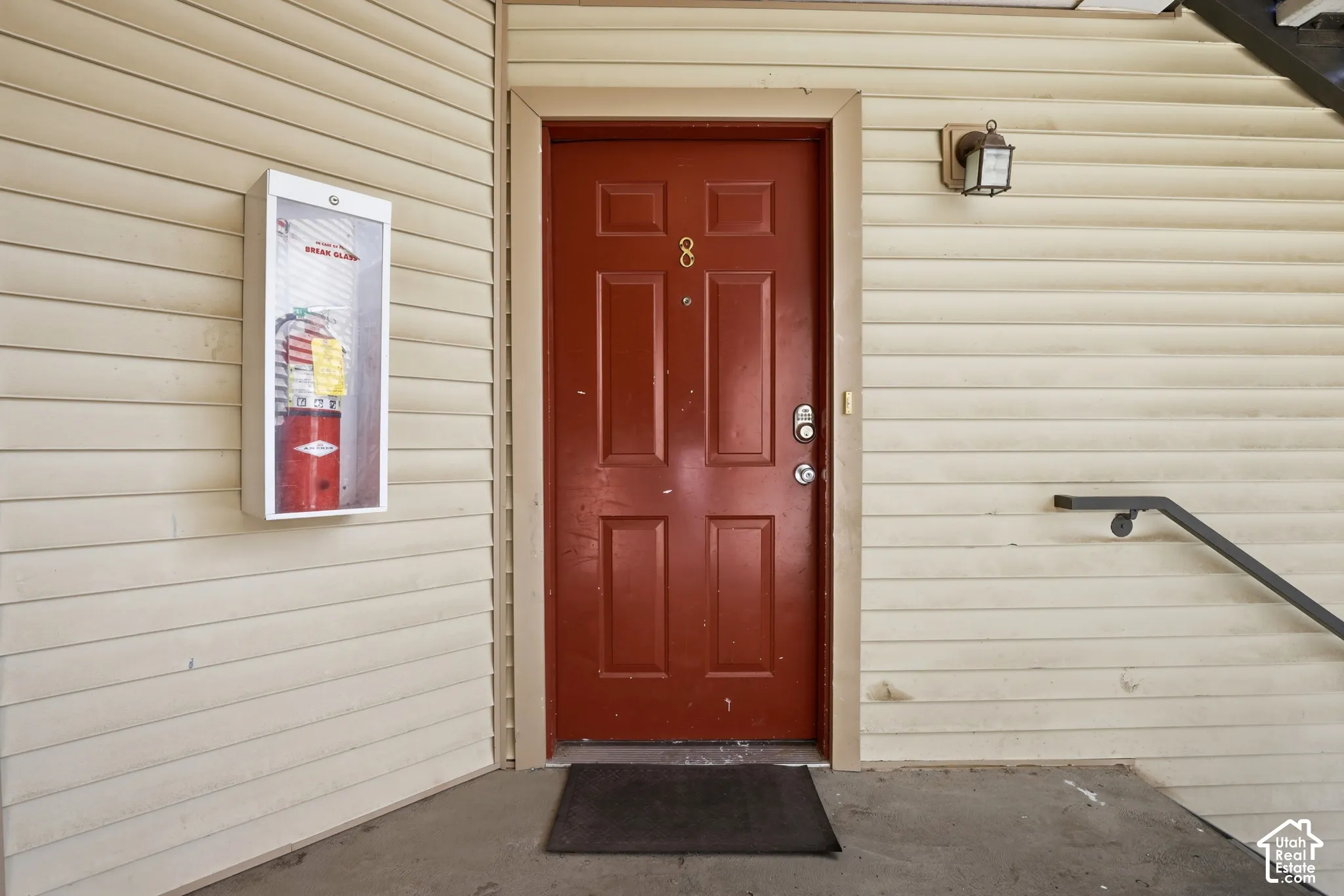 Exterior entryway featuring a red six-panel door, vinyl lap siding, wall-mounted lantern-style lighting, and a metal handrail
