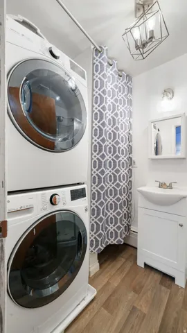 Laundry area with wood finished floors, stacked washer / dryer, a chandelier, and a baseboard radiator