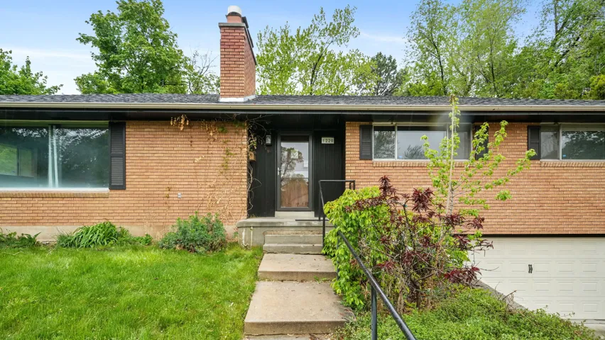 View of exterior entry with brick siding, a garage, a chimney, and a lawn