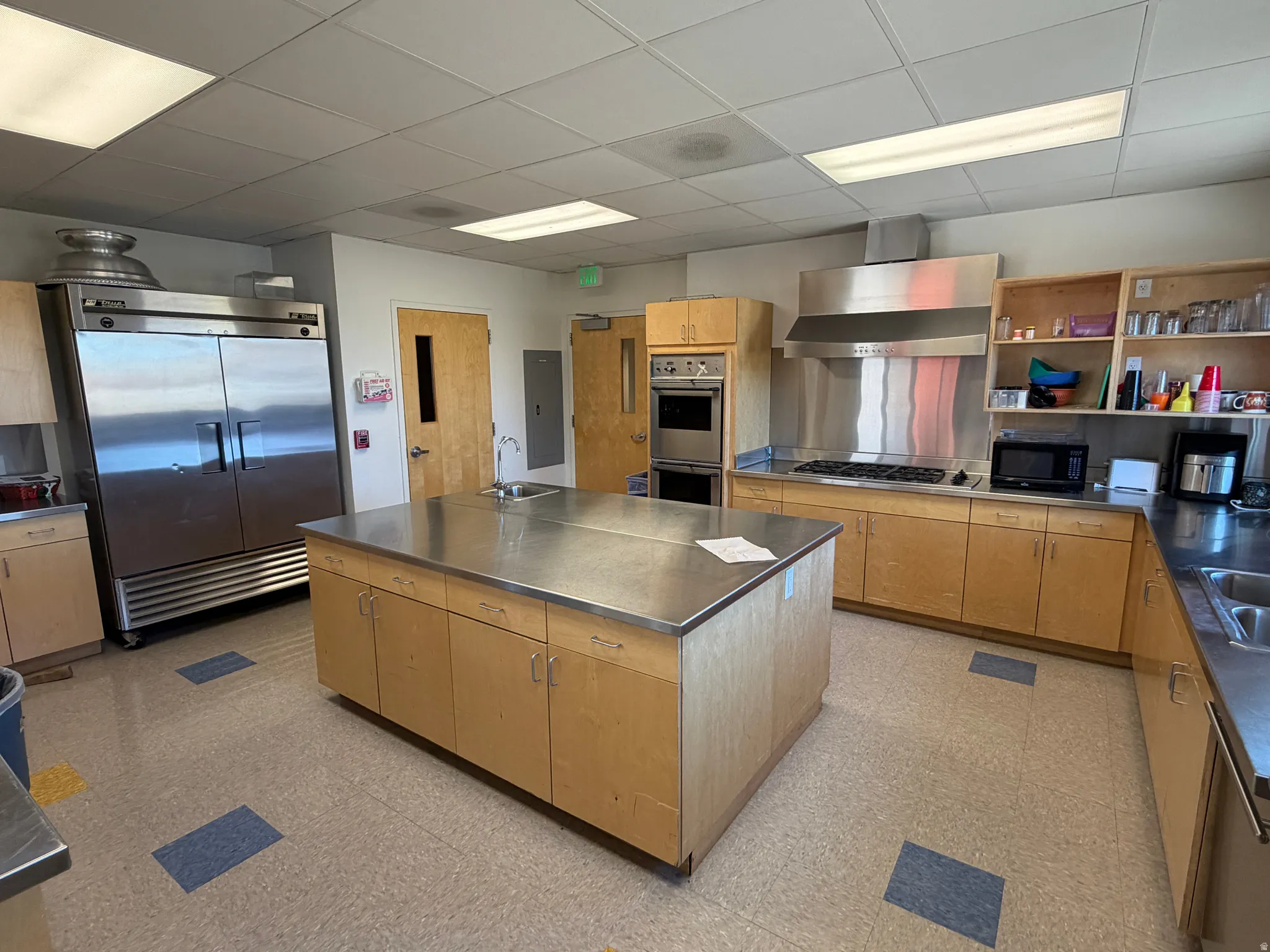 Kitchen with stainless steel counters, light wood finish cabinetry, stainless steel appliances, a drop ceiling, and an island with sink