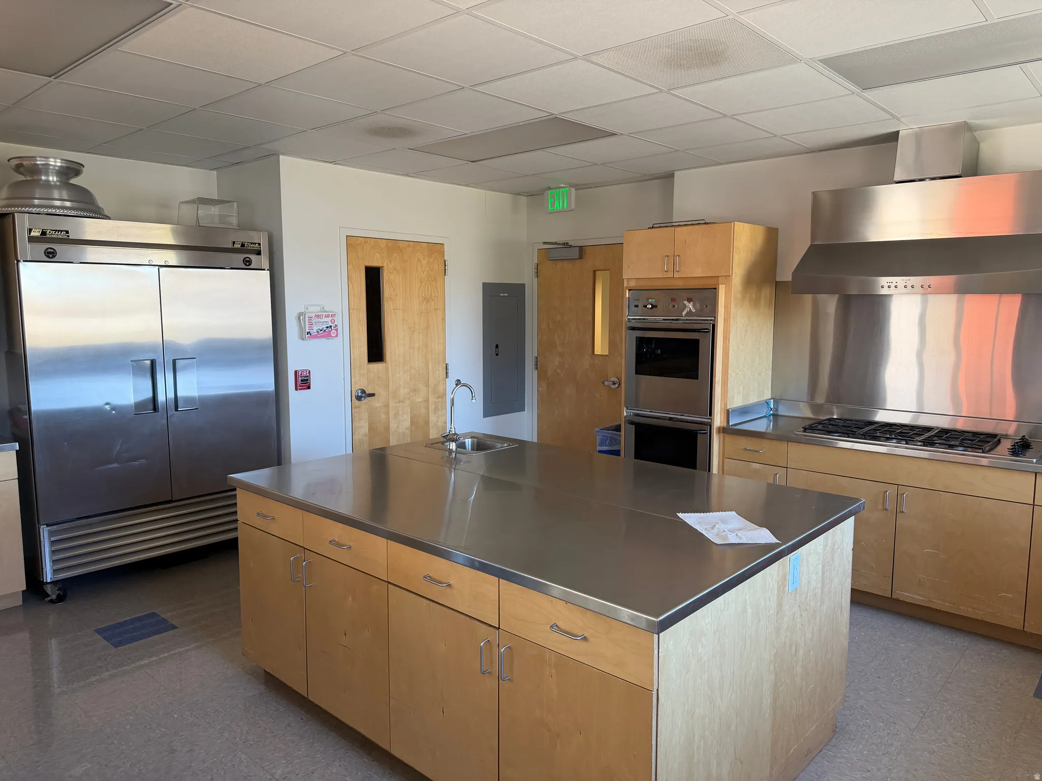Kitchen featuring stainless steel counters, stainless steel appliances, a large island, light wood finish cabinets, and a drop ceiling