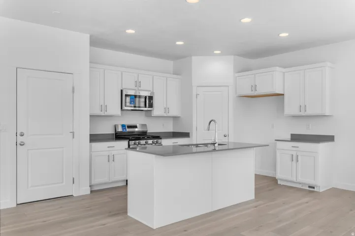 Modern kitchen featuring white cabinetry with brushed nickel hardware, stainless steel appliances, a center island with an integrated sink, dark countertops, and light wood-finish flooring