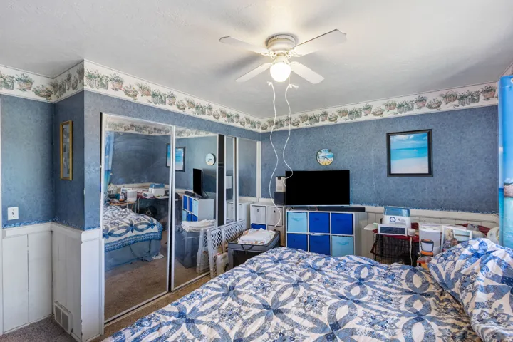 Bedroom featuring mirrored closet doors, a white ceiling fan with integrated lighting, textured blue wallpaper, and white wainscoting