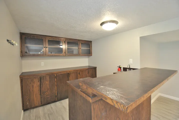 Indoor dry bar featuring light wood-type flooring, a textured ceiling, glass insert cabinets, and wood finish cabinets