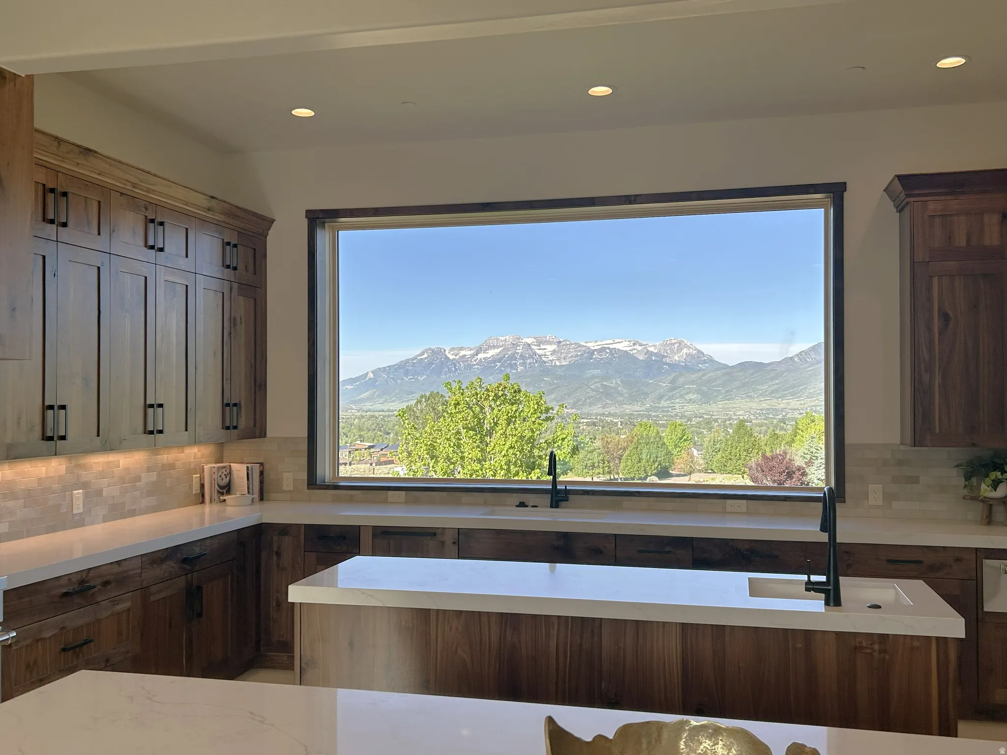 Kitchen featuring a large picture window with mountain and valley scenery, wood-finish cabinetry, light-toned countertops, tile backsplash, recessed lighting, and a kitchen island with a built-in sink
