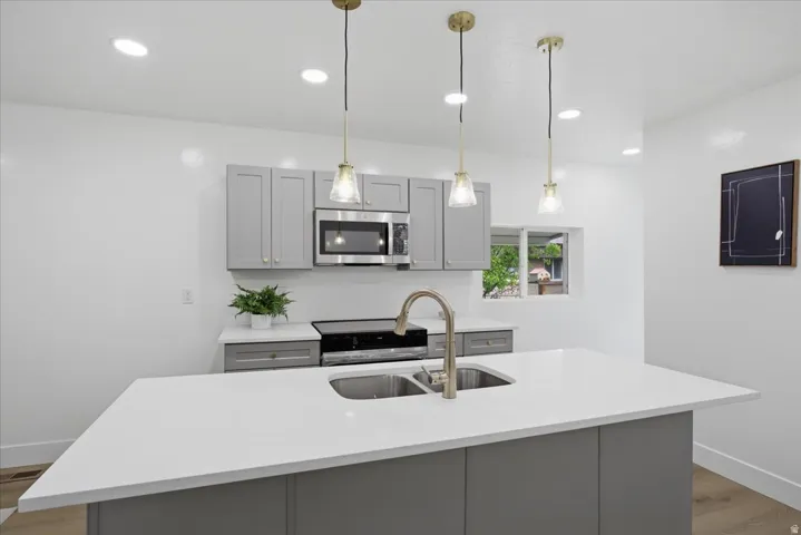 Kitchen with gray cabinetry, light wood finished floors, an island with sink, light stone countertops, and stainless steel microwave