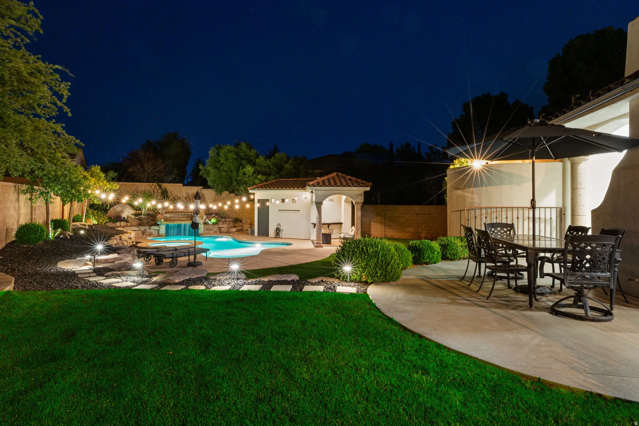 Expansive backyard featuring a custom-shaped pool with a waterfall, a built-in outdoor kitchen under a tiled roof, string lighting, manicured lawn, and a spacious concrete patio