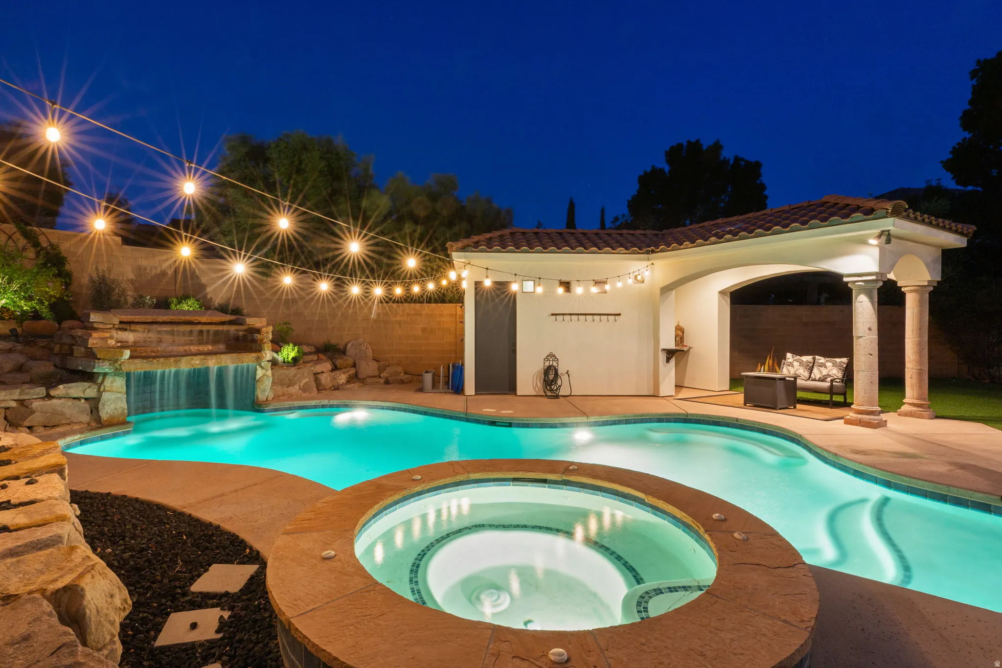 Resort-style backyard featuring a freeform swimming pool, integrated spa, rock waterfall feature, and covered patio with tiled roof
