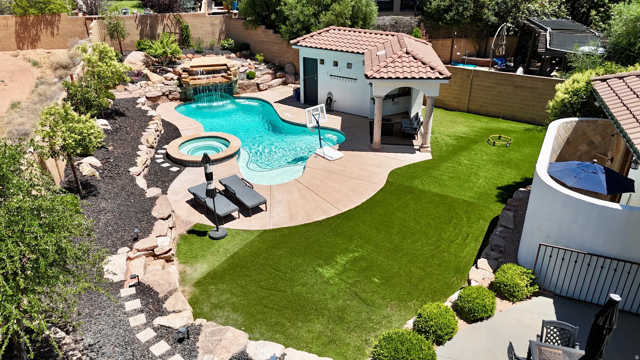 Backyard oasis featuring a freeform swimming pool with a waterfall, an integrated spa, a dedicated pool house with an attached covered patio, and a section of artificial turf