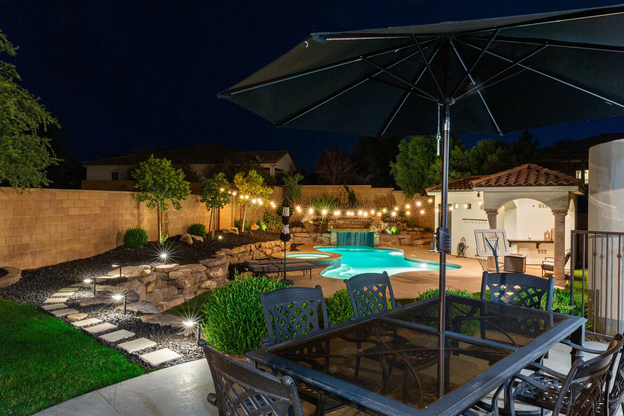 Resort-style backyard featuring a freeform swimming pool with a waterfall, an outdoor kitchen under a tiled pergola, and string lighting