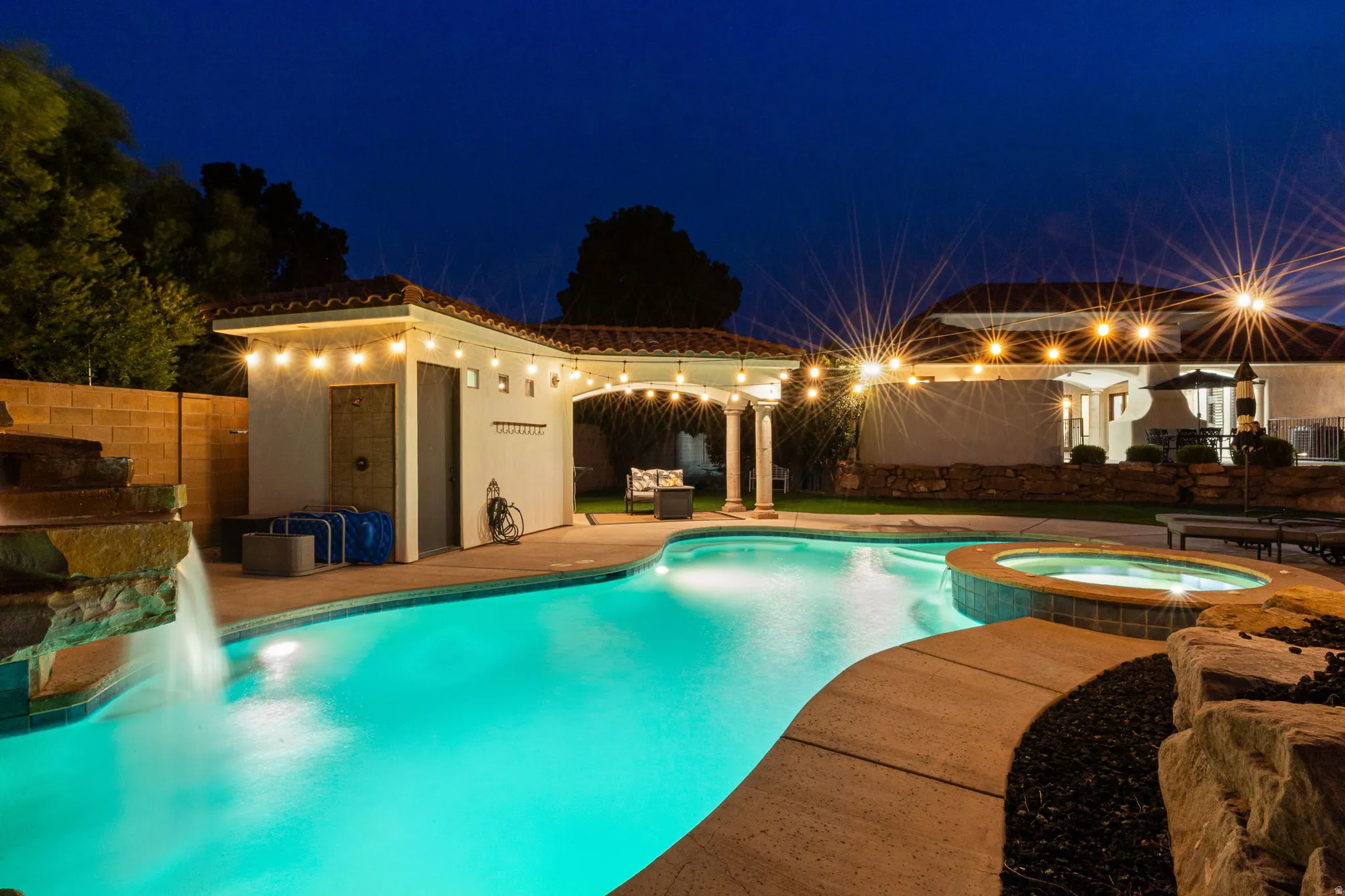 Custom-shaped swimming pool with an integrated spa, featuring a rock waterfall and perimeter lighting