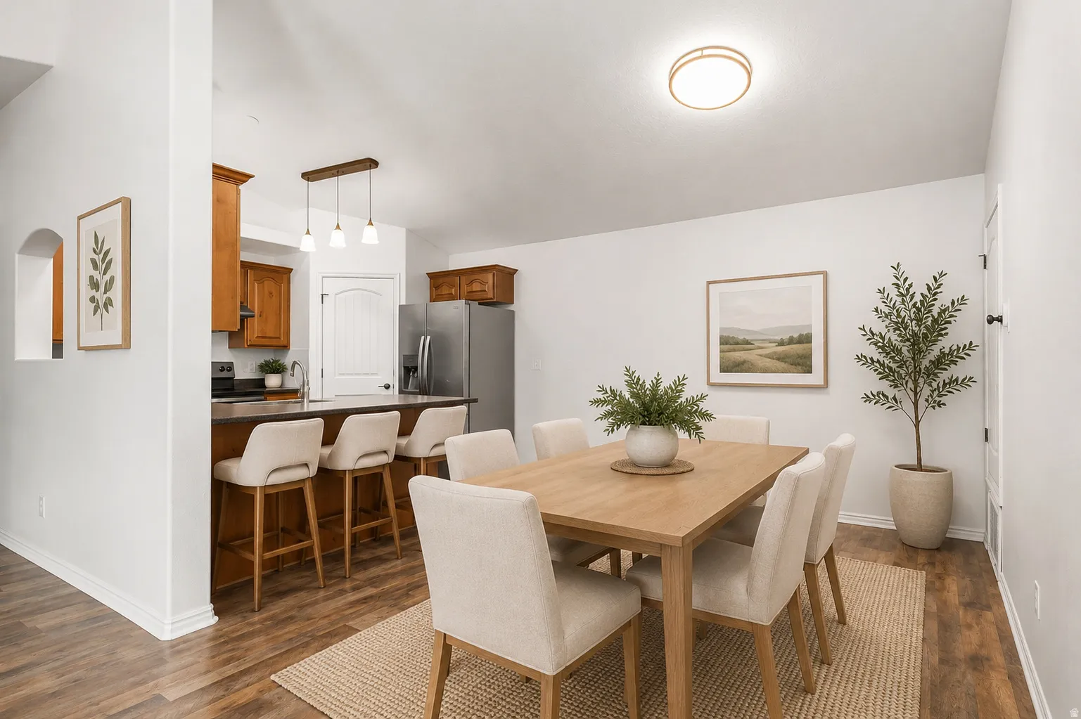 Open-concept dining and kitchen area featuring a wood-finish dining table with upholstered chairs, warm wood cabinetry with stainless steel appliances, pendant lighting, recessed ceiling fixtures, and wood-finish flooring throughout