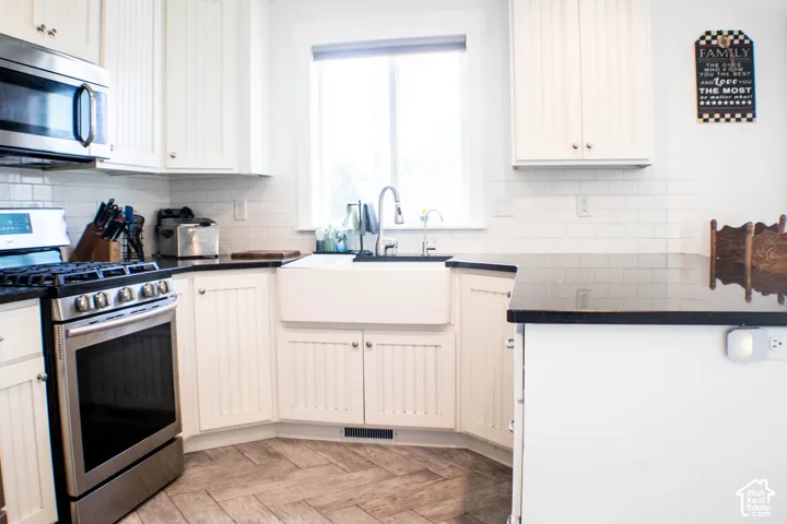 White beadboard cabinetry, stainless steel appliances, white subway tile backsplash, farmhouse-style sink, and dark countertops