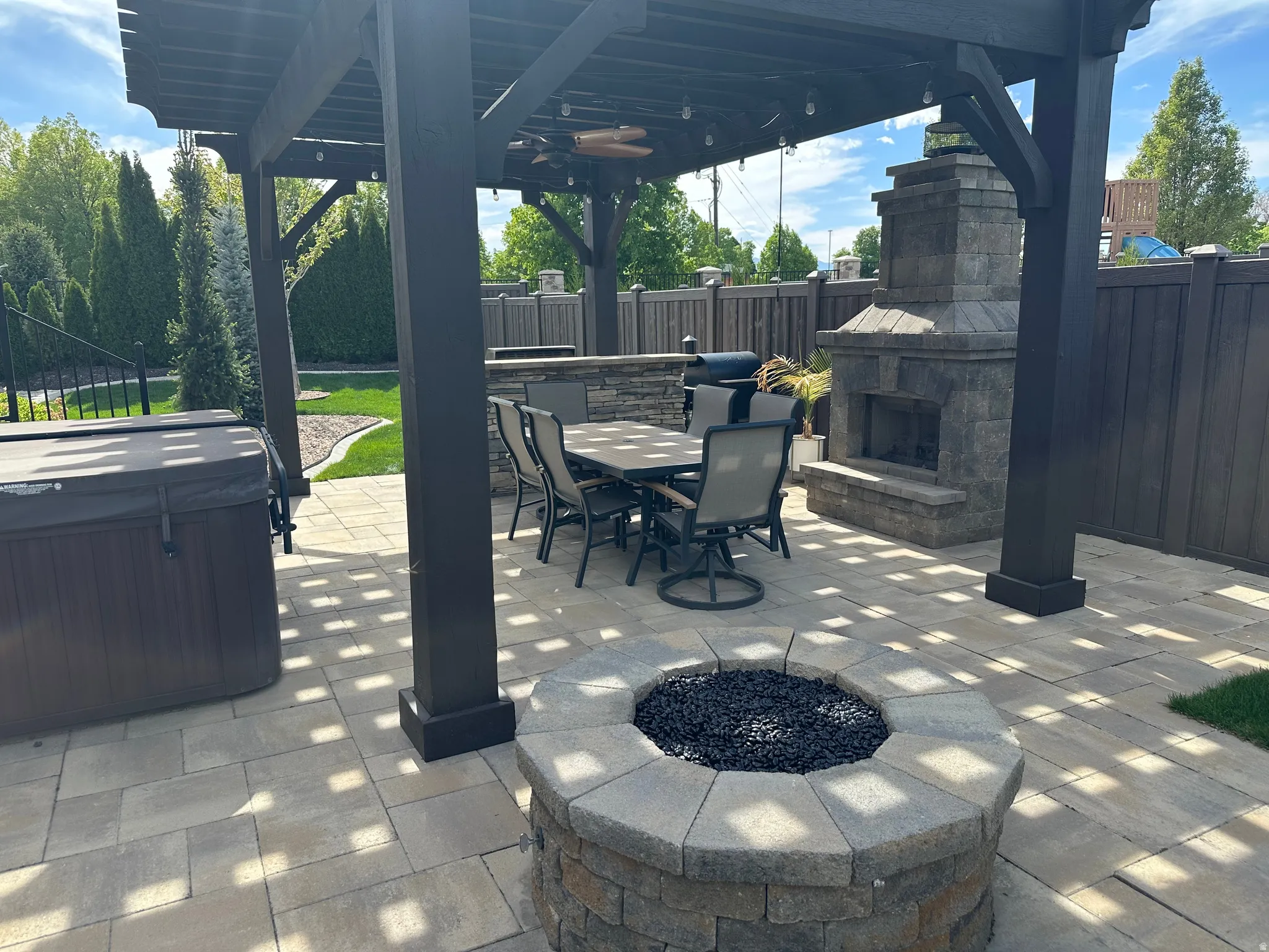 Fenced backyard with a hot tub, an outdoor stone fireplace, a patio area, outdoor dining area, and a pergola