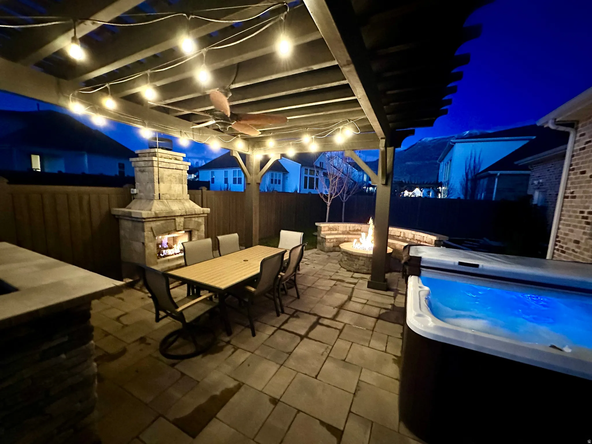 Patio at night featuring outdoor dining area, a patio area, a fire pit, a fenced backyard, and an outdoor stone fireplace