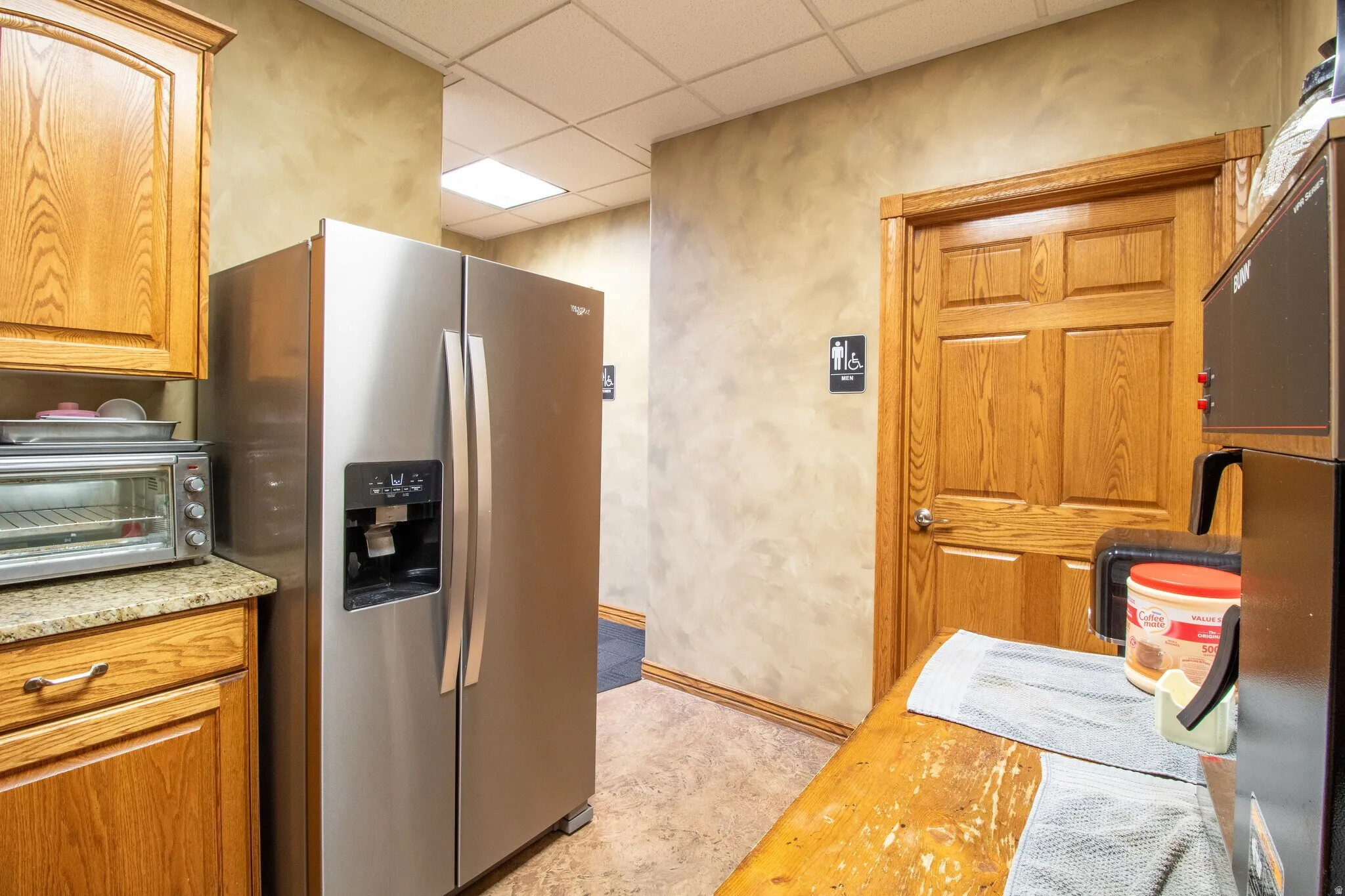 Kitchen with a paneled ceiling, stainless steel fridge with ice dispenser, wood finish cabinets, and light stone countertops