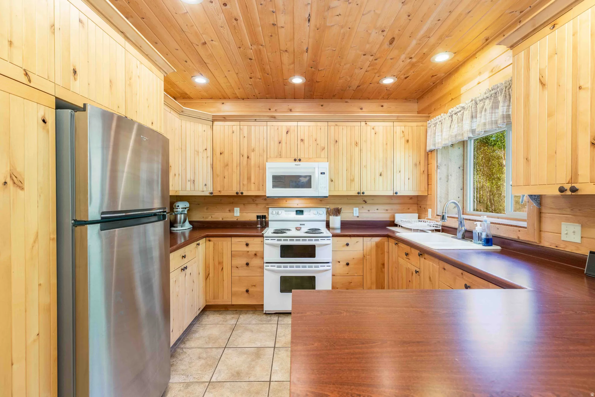 Kitchen featuring white appliances, dark countertops, light wood finish cabinetry, recessed lighting, and light tile patterned floors