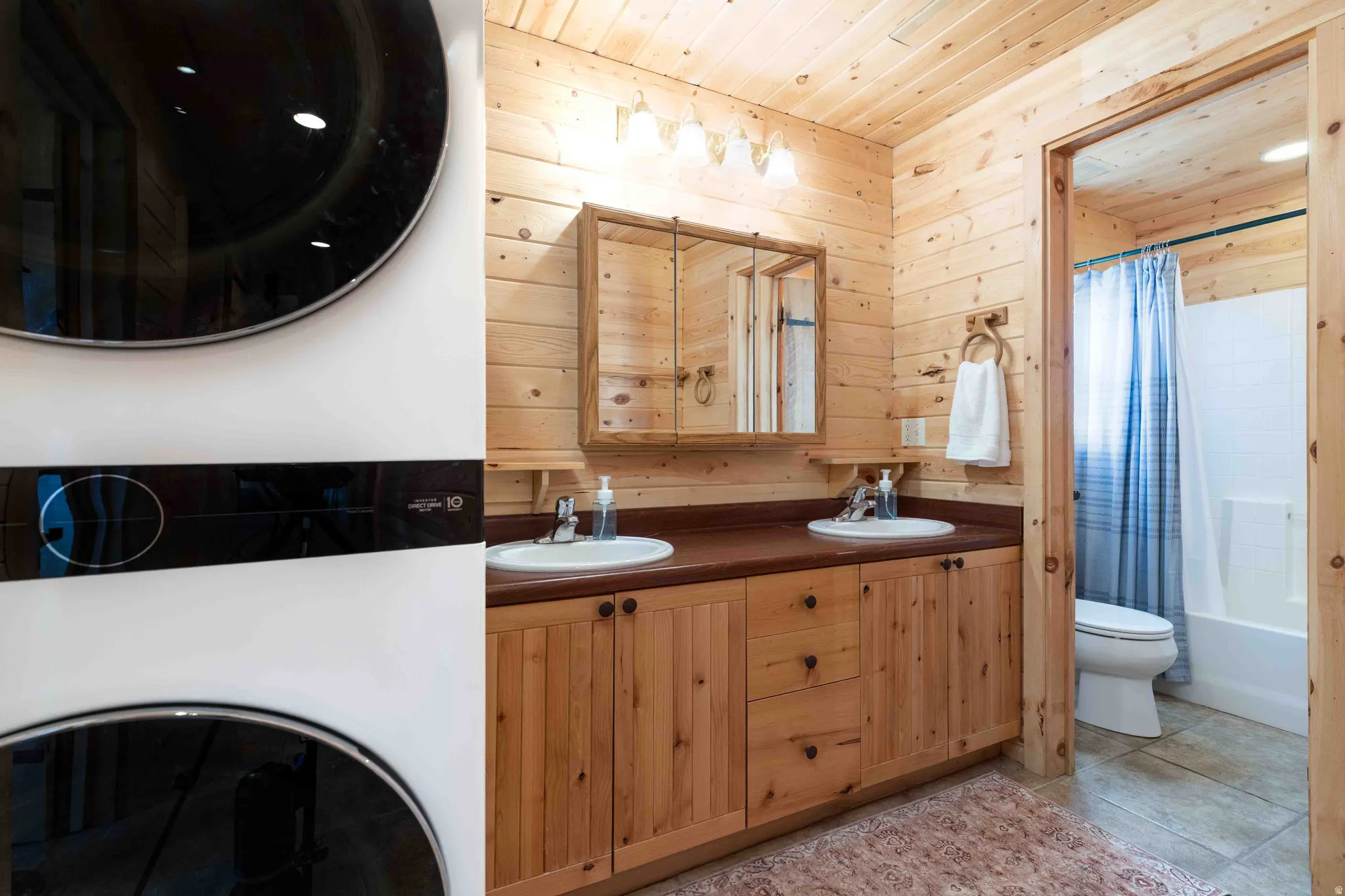Full bathroom featuring double vanity, wood walls, stacked washer / drying machine, shower / bath combo with shower curtain, and wood ceiling