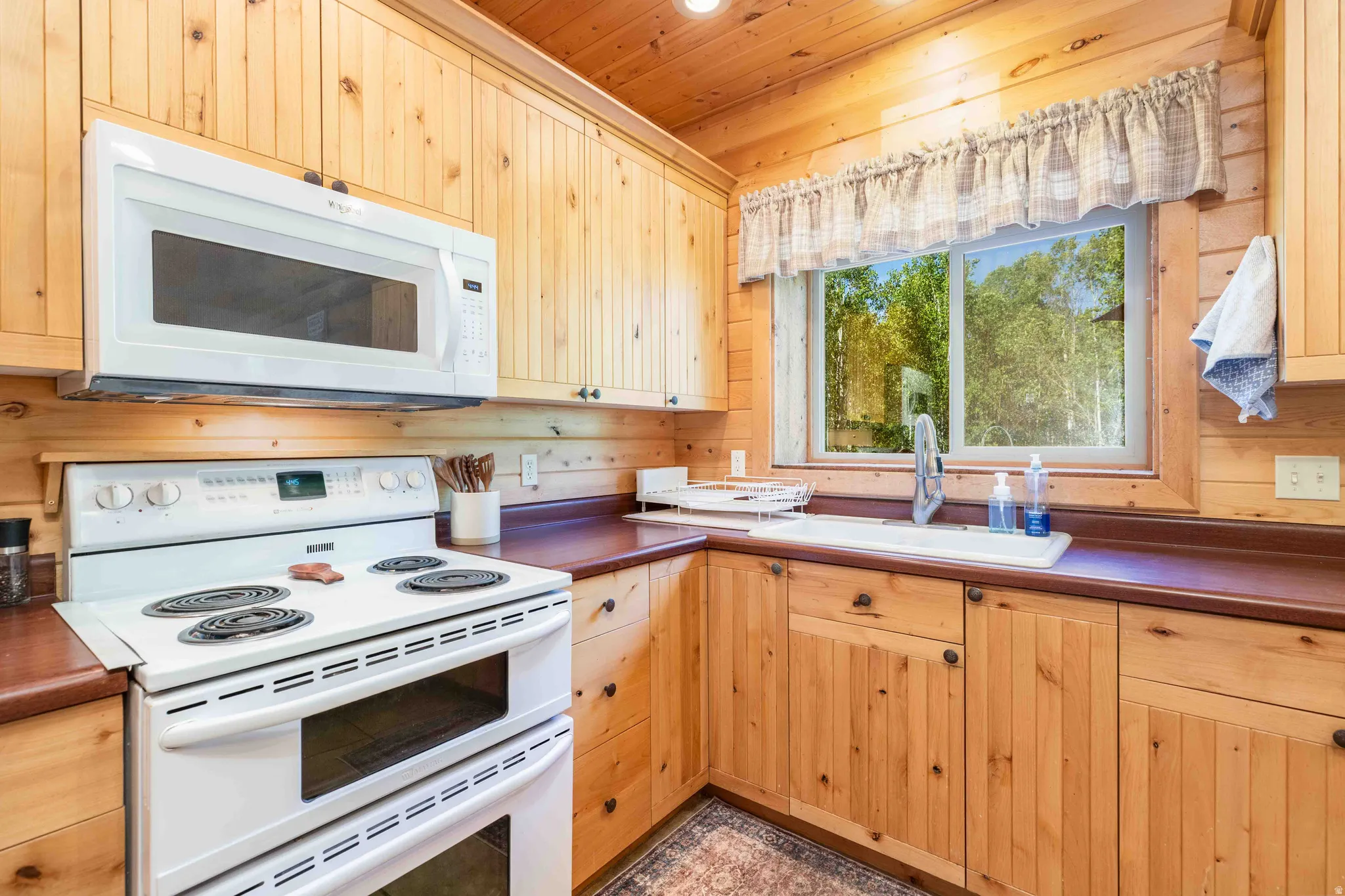 Kitchen featuring white appliances, dark countertops, wood walls, light wood finish cabinetry, and wood ceiling