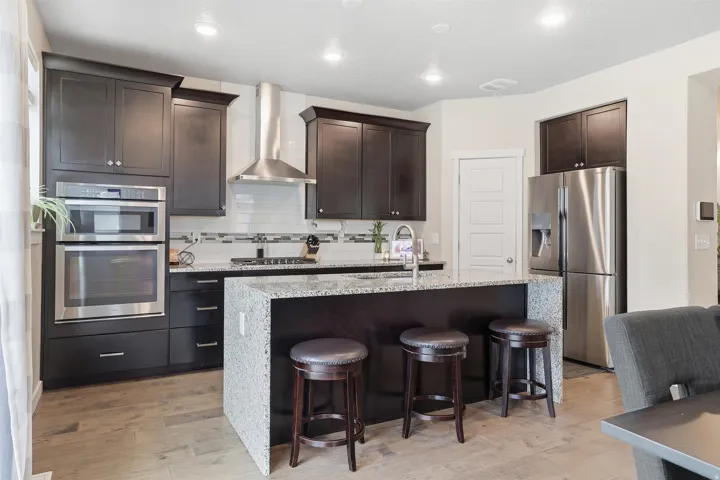 Kitchen with light stone counters, stainless steel appliances, a kitchen island with sink, dark wood finish cabinets, and recessed lighting