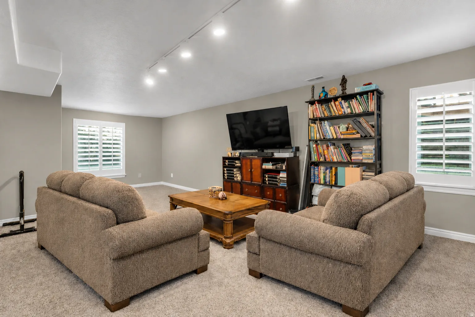 Recessed track lighting, two windows with plantation shutters, light-toned carpeting, and a built-in media console
