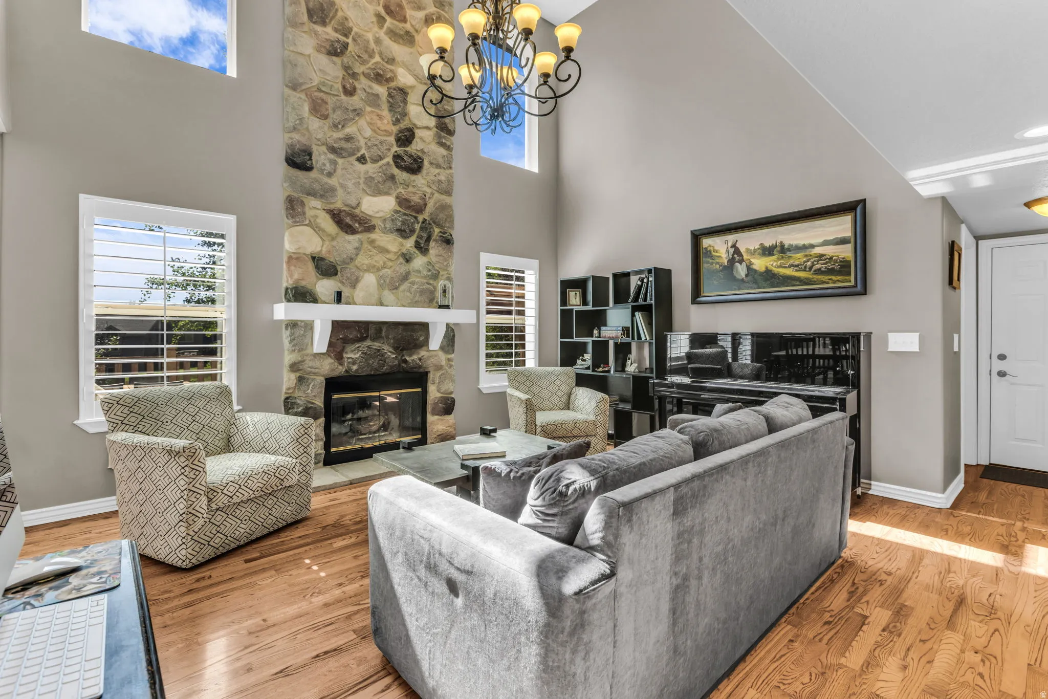 Spacious living area featuring a two-story stone fireplace with decorative mantel, hardwood flooring throughout, recessed lighting, built-in shelving with display cabinetry, large windows with plantation shutters, and a decorative chandelier