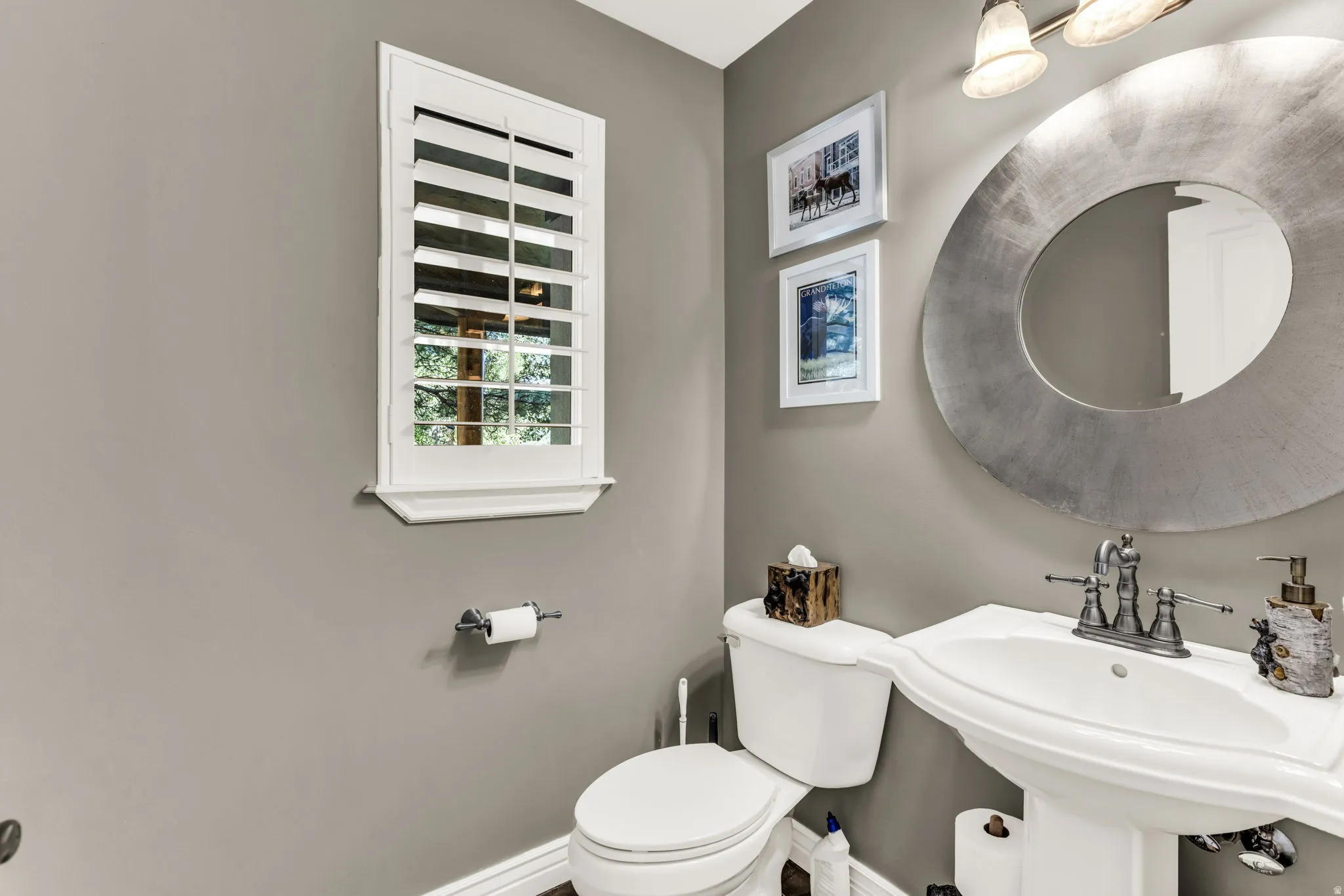 Pedestal sink with dual-handle faucet, circular wall mirror, and window with plantation shutters