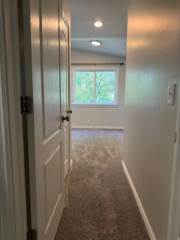 Carpeted hallway featuring a window with exterior tree scenery, white trim, and a vaulted ceiling with recessed and flush-mount lighting