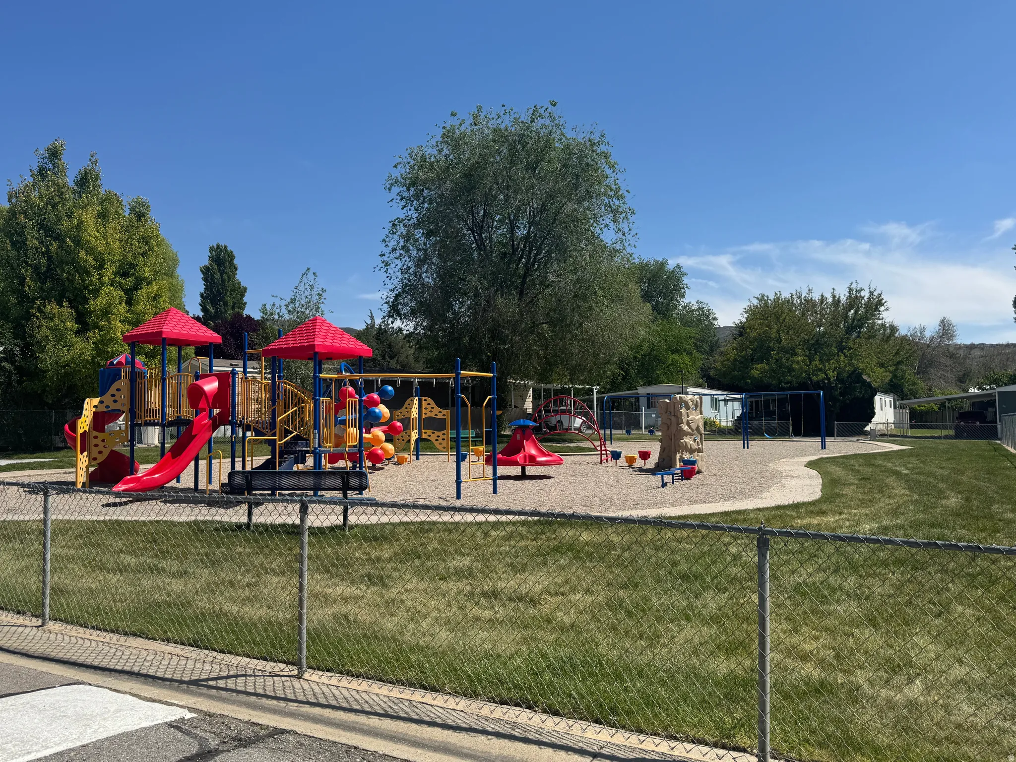 Community playground featuring a multi-level play structure with red slides and colorful accents, a rock climbing feature, and a swing set
